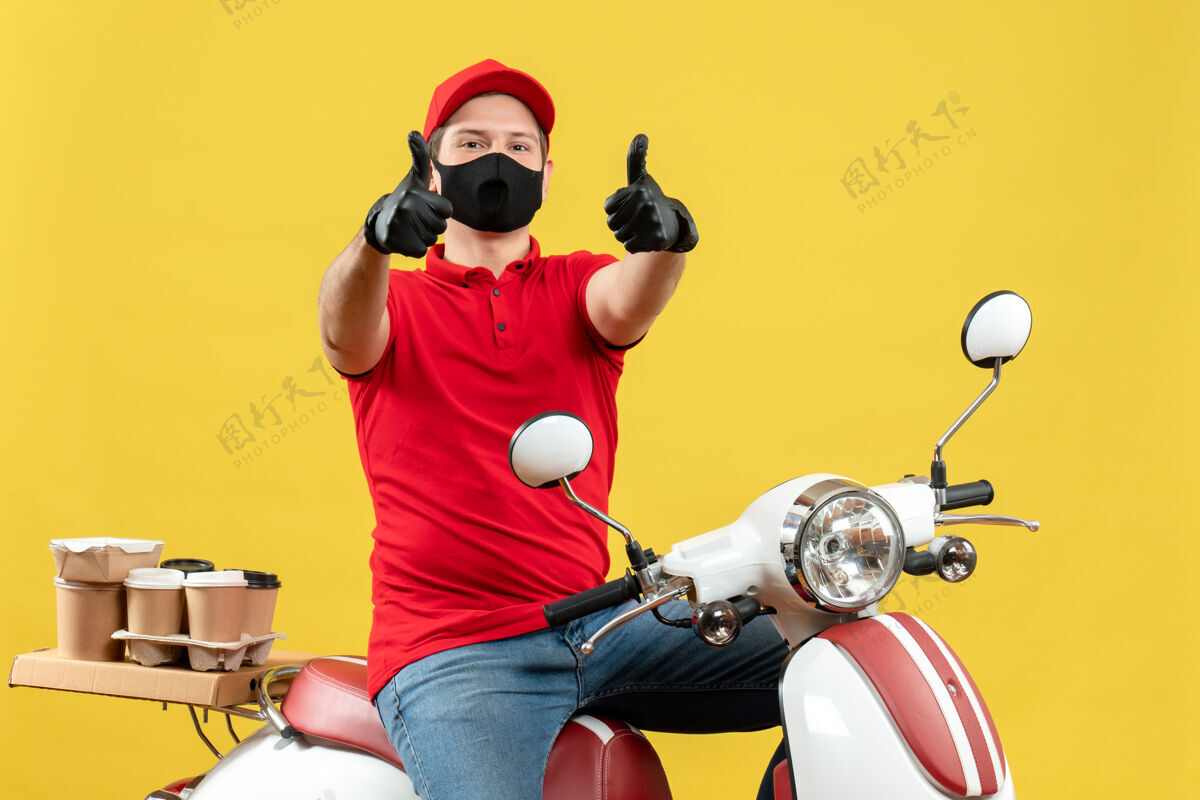 好的戴着医用面罩 身穿红色上衣 戴着帽子手套 面带微笑的年轻人坐在踏板车上 在黄色背景上做着“ok”的手势面罩手势坐着