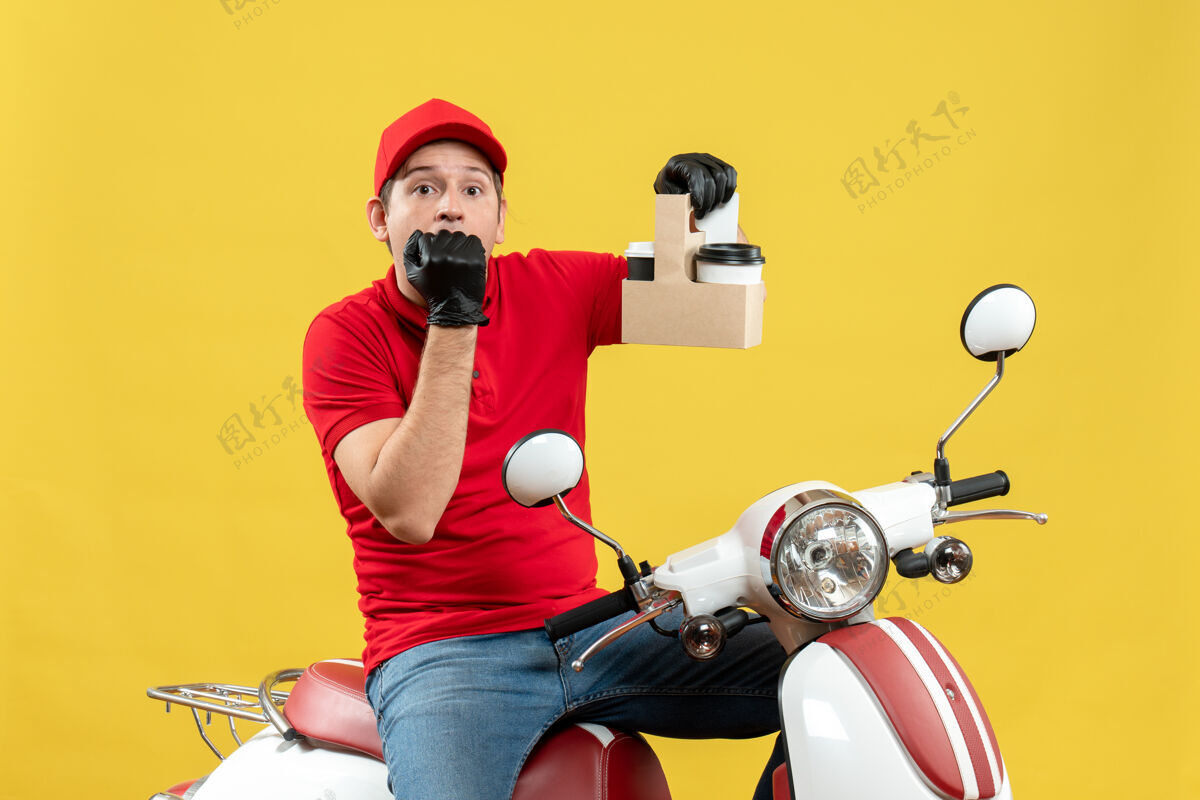 交付前视图混乱的快递员身穿红色上衣 戴着帽子手套 戴着医用面罩 坐在滑板车上拿着订单坐摩托车摩托车