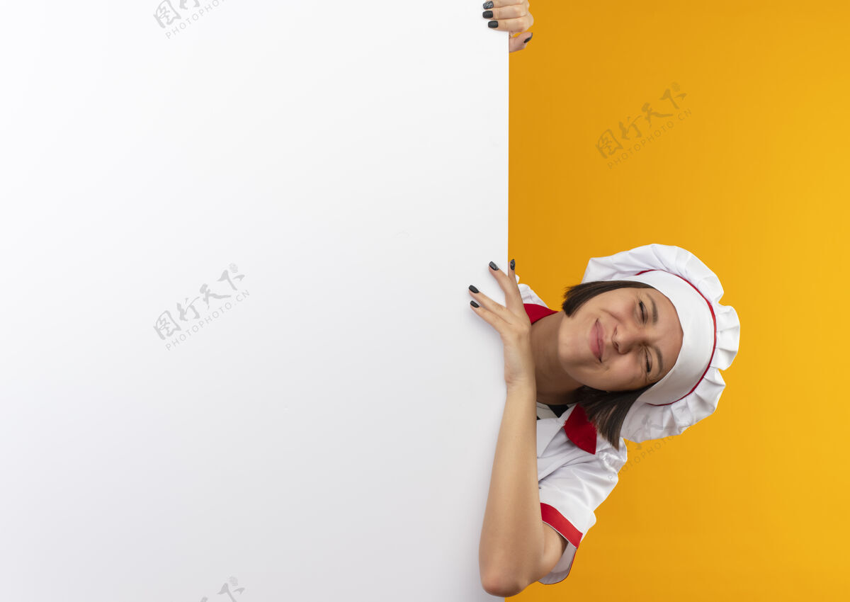 女身着厨师制服的年轻女厨师站在白色的墙壁后面 手握并把手放在墙上 闭着眼睛 孤立地站在橙色的墙上封闭请白