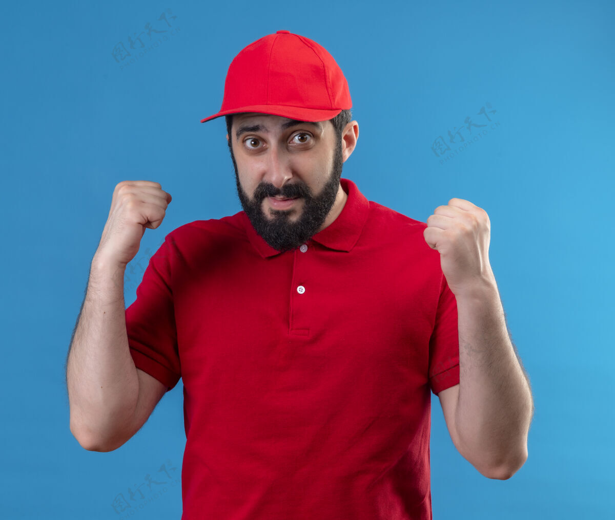 帅哥年轻帅气的送货员穿着红色制服 举着高帽的拳头孤立在蓝色的墙上表情人帽子