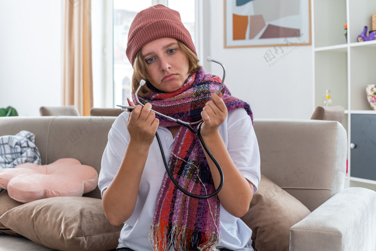 不健康不健康的年轻女子戴着帽子 脖子上围着暖和的围巾 感觉不舒服 生病 患感冒和流感 用听诊器听脉搏 坐在明亮的客厅里的沙发上看起来很担心听诊器脉搏脖子