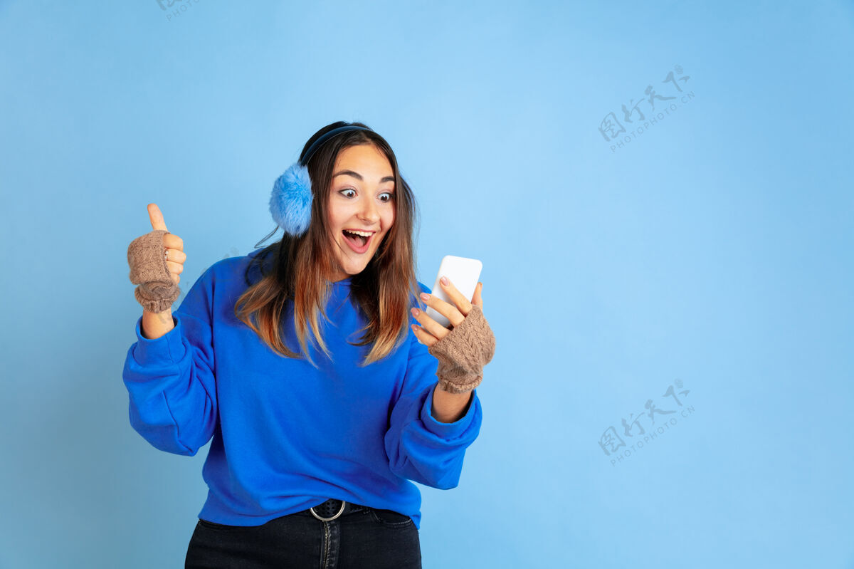 员工使用电话 快乐蓝色工作室背景上的白种女人肖像穿着暖和衣服的漂亮女模特人类情感的概念 面部表情 销售 广告冬天的心情 假期成功工作一半