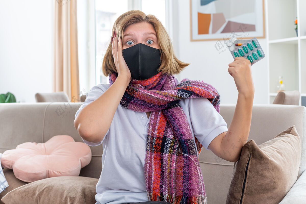 坐着不健康的年轻女子 脖子上围着暖和的围巾 戴着面膜 感觉不舒服 生病 患流感 吃感冒药 坐在客厅的沙发上 看起来很困惑感冒不适生病