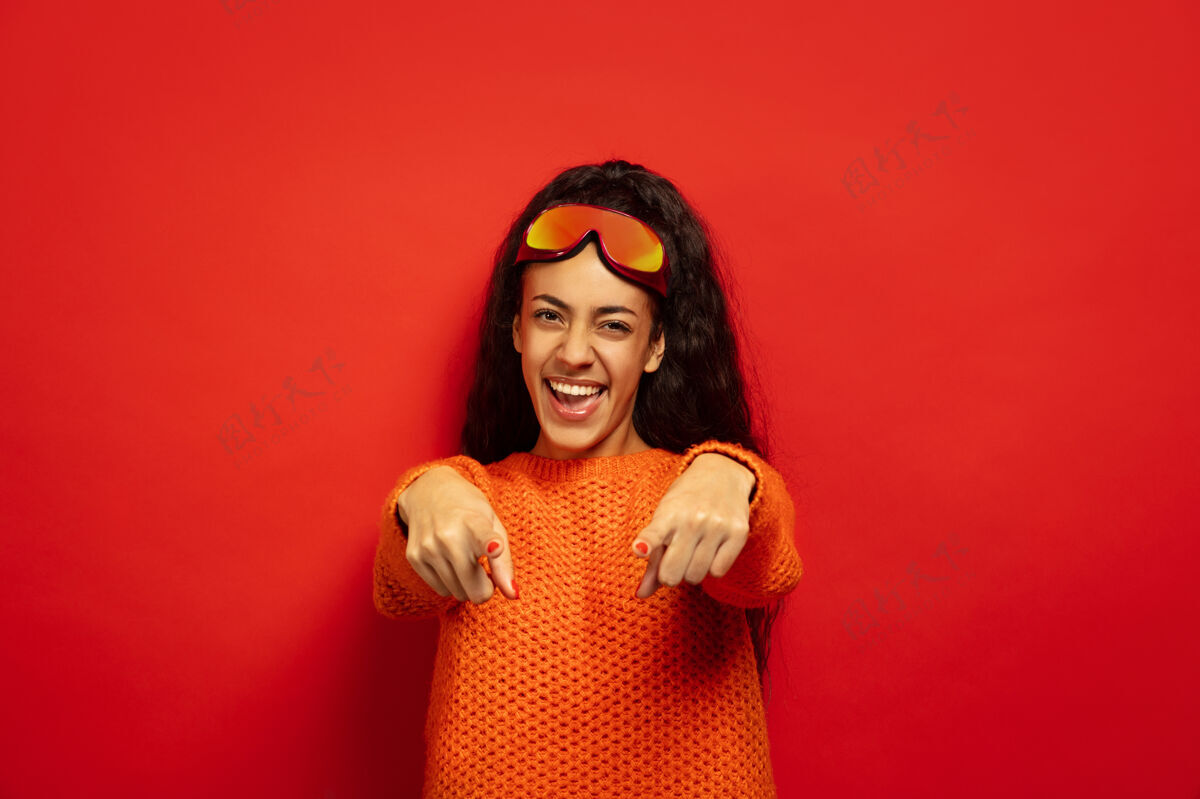 惊喜美国黑人年轻黑发女子的肖像滑雪面具上的红色工作室背景概念的人类情感 面部表情 销售 广告 冬季运动和假期指着 笑手长表情