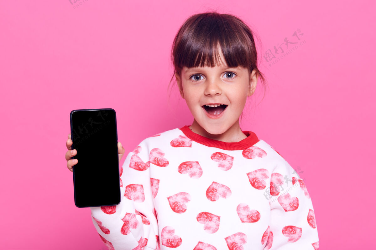 在线惊讶的小女孩拿着智能手机 一脸惊讶的表情 张开嘴 隔着粉色的墙壁摆出孤立的姿势博主联系人大笑