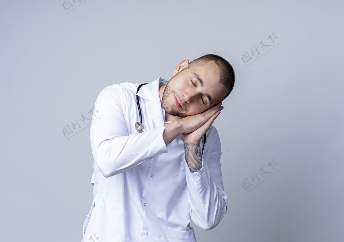 手势昏昏欲睡的年轻男医生 穿着医用长袍 脖子上戴着听诊器 在白墙上做着与世隔绝的睡姿人年轻睡眠