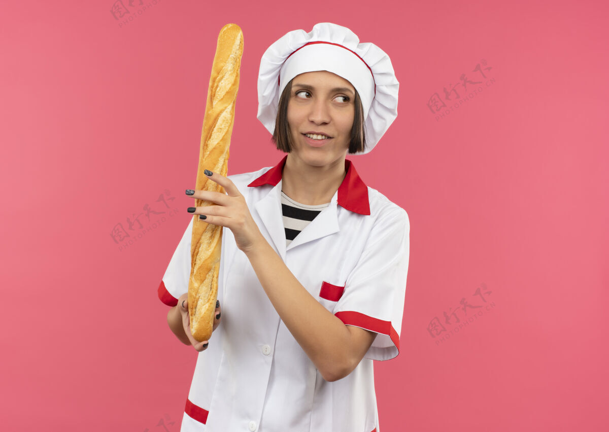 厨师面带微笑的年轻女厨师身着厨师制服拿着面包棒看着隔离在粉红色墙上的一面姿势表情制服