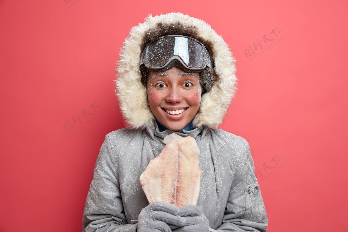 毛皮冰雪覆盖的快乐北极女人笑容满面 穿着带兜帽的外套 戴着温暖的手套 在冬天滑雪钓鱼或滑雪板滑雪后高兴地抱着冻鱼积极的休息爱好概念冬天欢呼正面