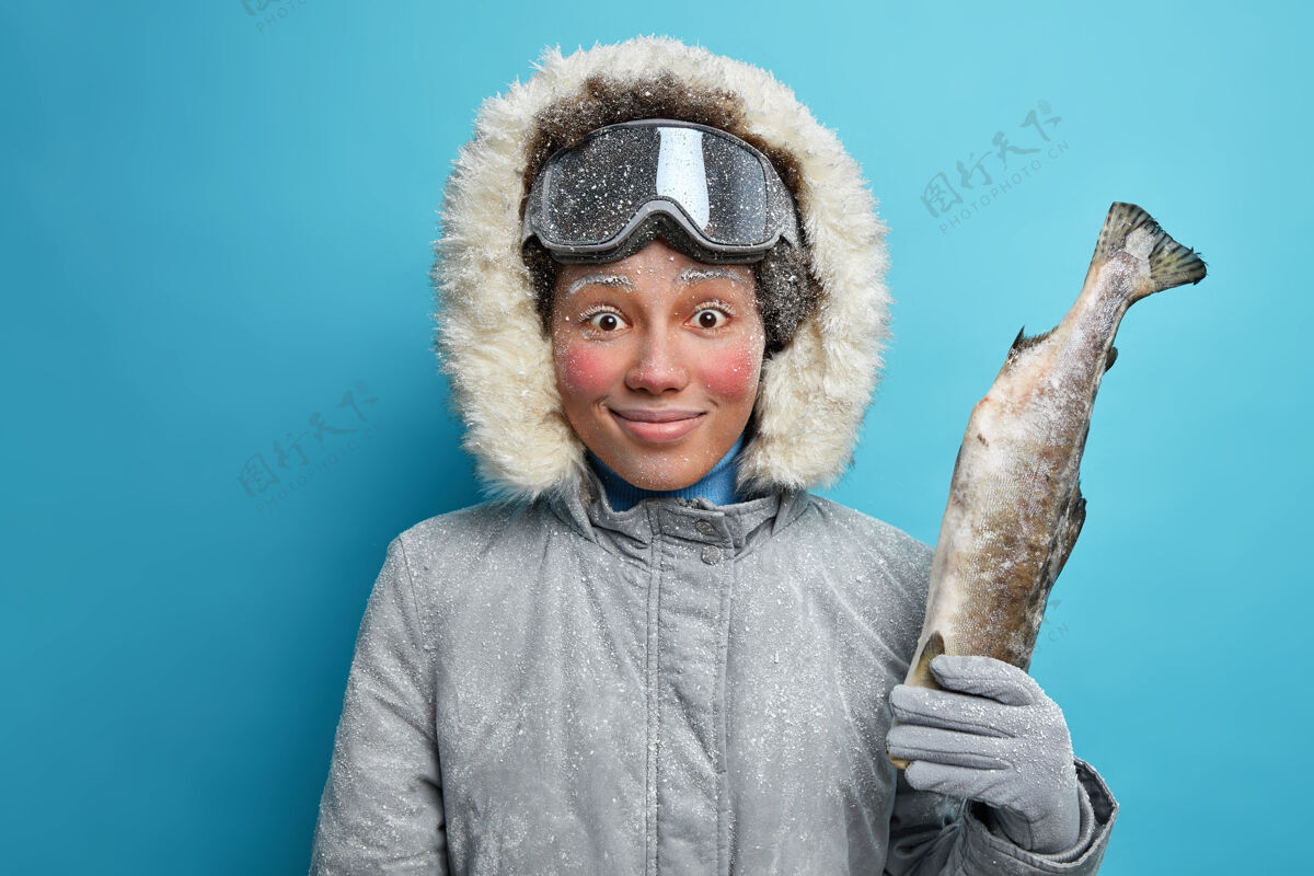 民族高兴的女人愉快地享受冬天钓鱼的微笑 抱着大钓到的鱼看起来很生气 穿着灰色夹克 带着兜帽 在寒冷的冬天有空 天气冻红了脸女人冬天蓝色