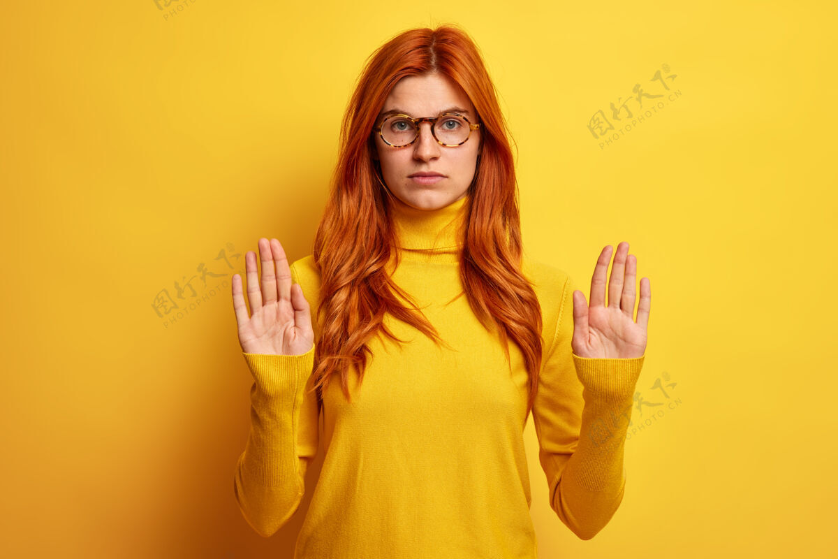 禁止严肃的年轻红发女子照片显示停车手势做禁牌拉手掌 戴眼镜和高领毛衣女人防御成人