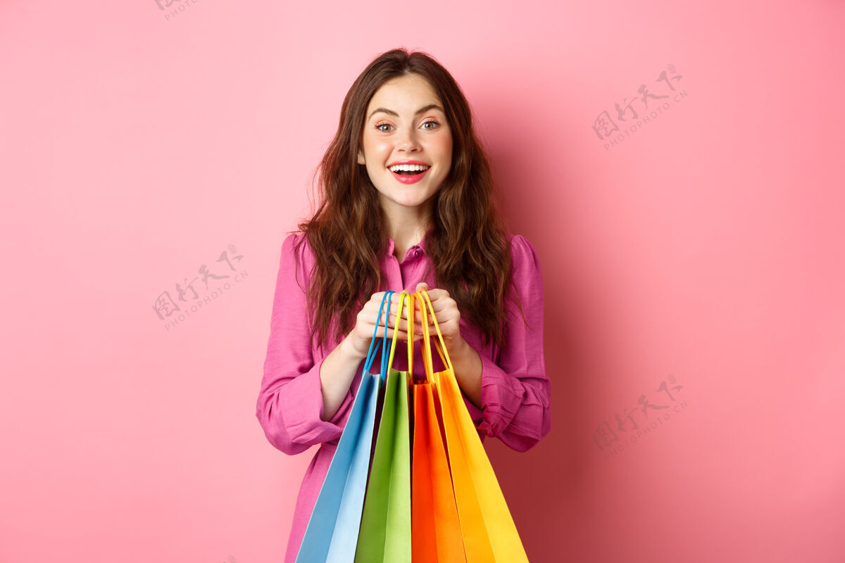 请年轻漂亮的女士拿着购物袋 兴奋地微笑着 打折购物 站在粉红色的墙上时尚积极欢呼