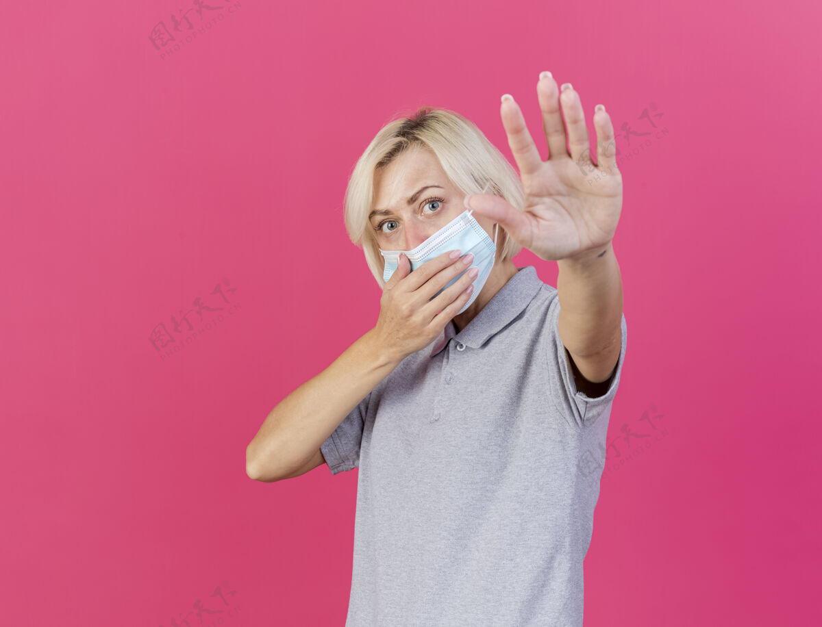 站自信的年轻金发病斯拉夫妇女戴着医用口罩 把手放在嘴上 举起手站在粉红色的墙上 有复制空间养年轻疾病