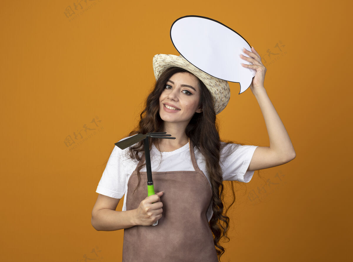 演讲微笑的年轻女园丁穿着制服 戴着园艺帽 拿着演讲泡泡和锄头耙 隔离在橙色的墙上 留着复制空间持有园艺花园