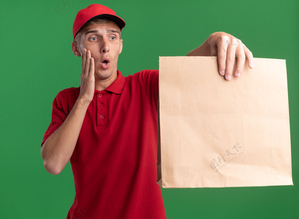 焦虑焦急的年轻金发送货男孩把手放在脸上拿着 看着绿色的纸包装交货手绿