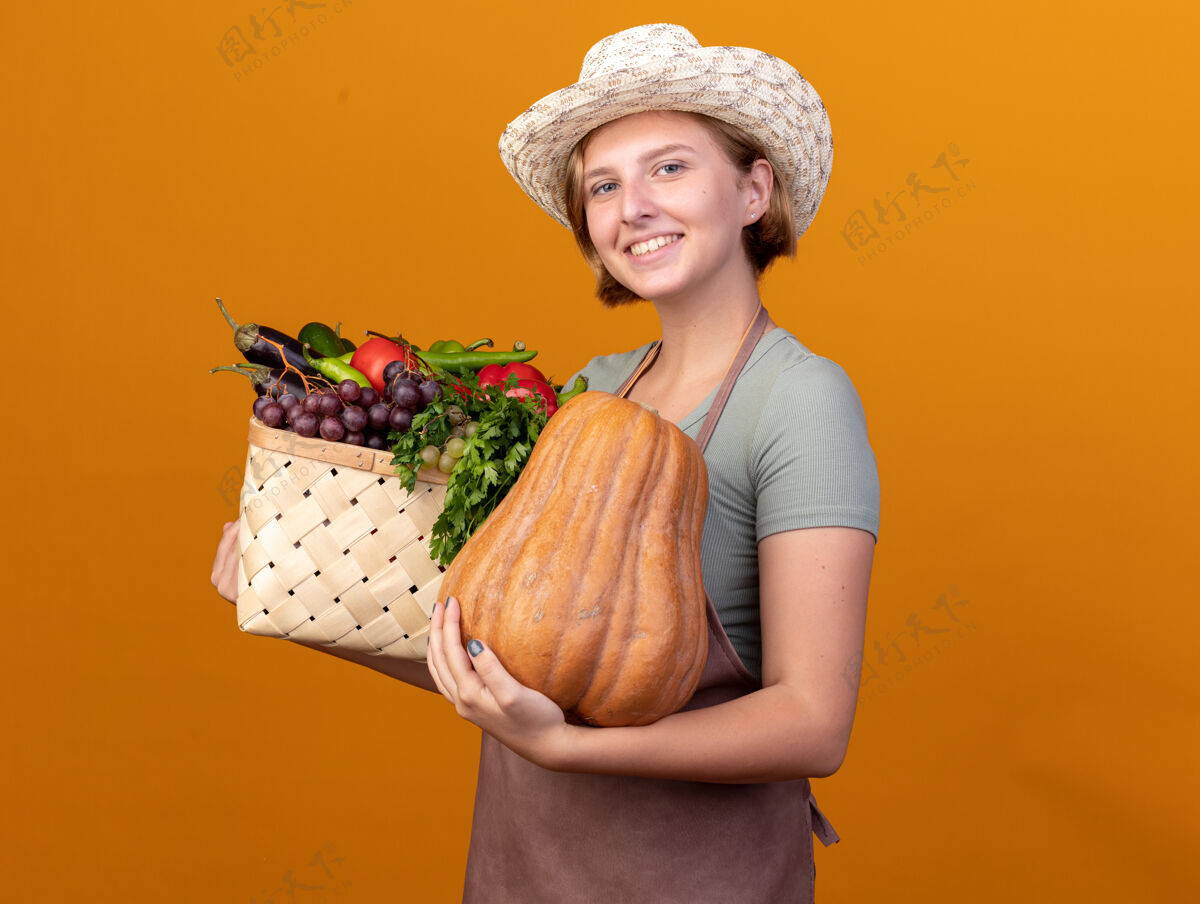 雌微笑着的年轻的斯拉夫女园丁戴着园艺帽 手里拿着蔬菜篮子和橘子上的南瓜南瓜篮子帽子