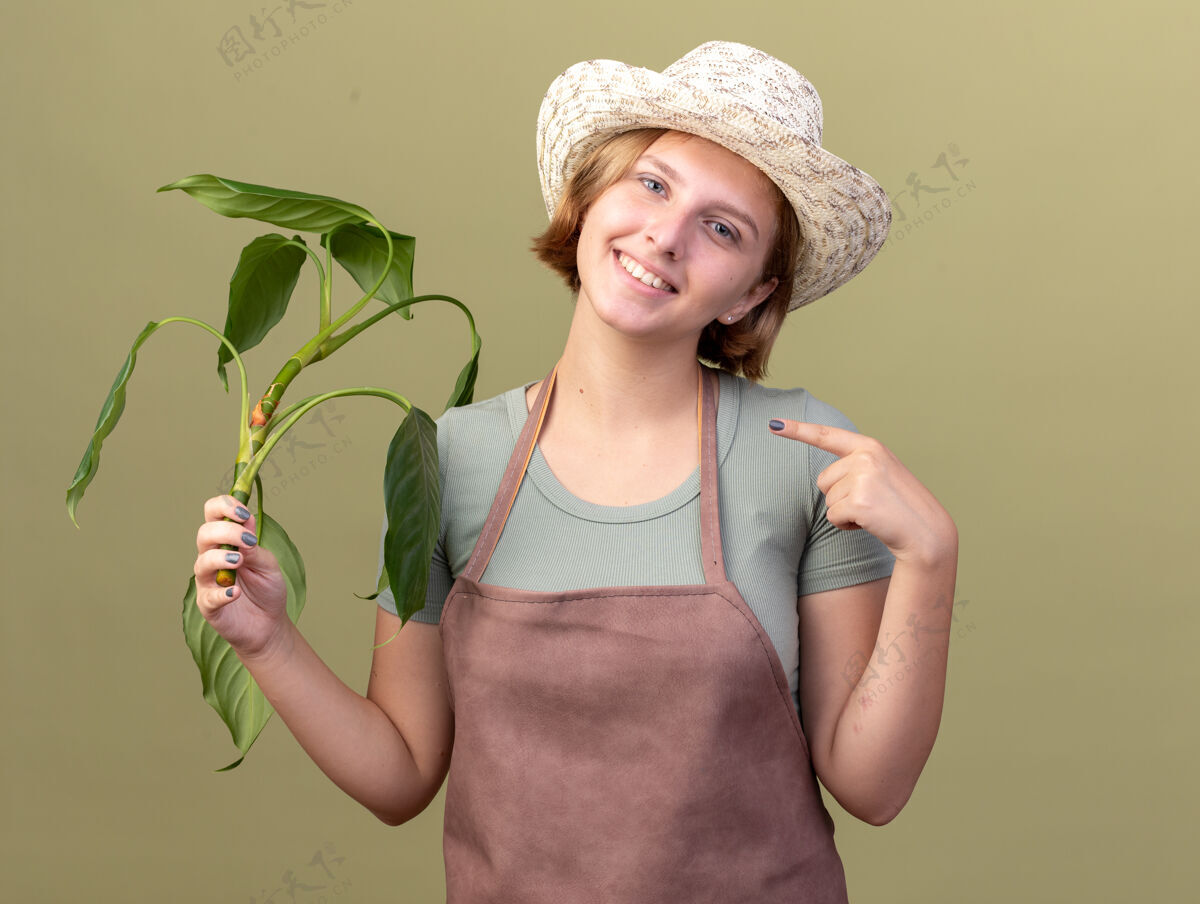 指点微笑着的年轻的斯拉夫女园丁戴着园艺帽 手持并指着橄榄绿上的植物帽子斯拉夫女