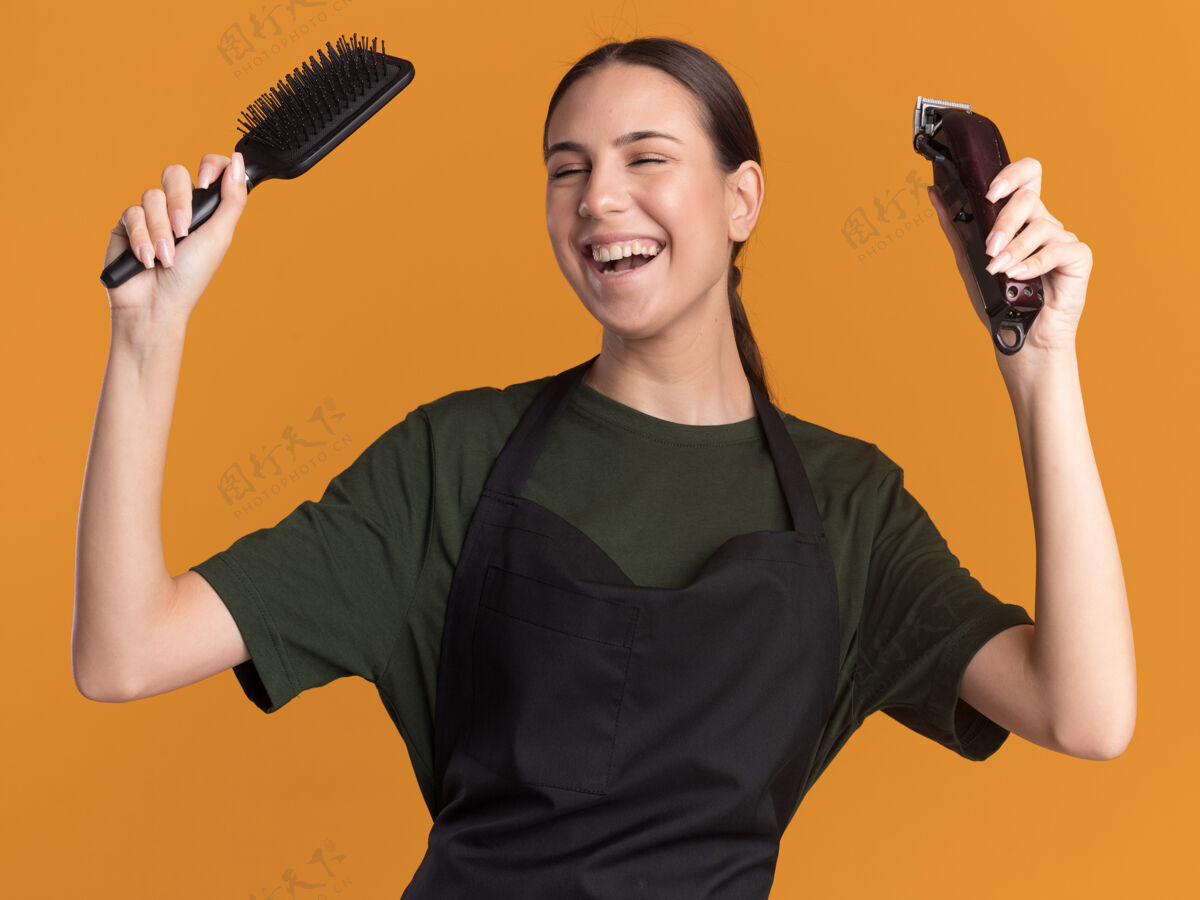 梳子快乐的年轻黑发女理发师在制服拿着剪子和梳子在橙色拿着橙色年轻人