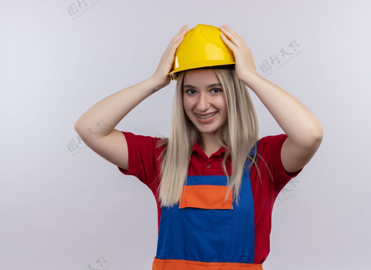 女孩微笑着的年轻金发工程师建筑工女孩 穿着制服 戴着牙套 手戴着安全帽 站在隔离的白色空间里微笑工程师建筑工人
