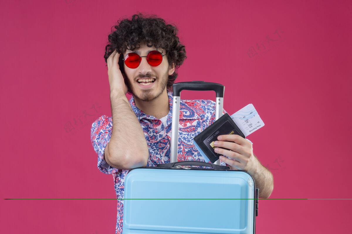 手高兴的年轻英俊的卷发旅行者男子戴着墨镜拿着机票 钱包把手放在头上和手提箱上闭着眼睛在孤立的粉红色空间与复印空间飞机眼睛持有
