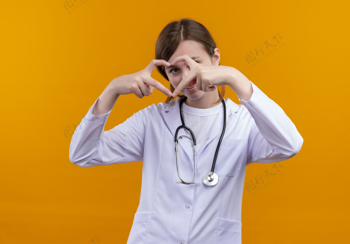 女性高兴的年轻女医生穿着医用长袍和听诊器做心脏的迹象 并通过它看孤立的橙色空间年轻听诊器穿