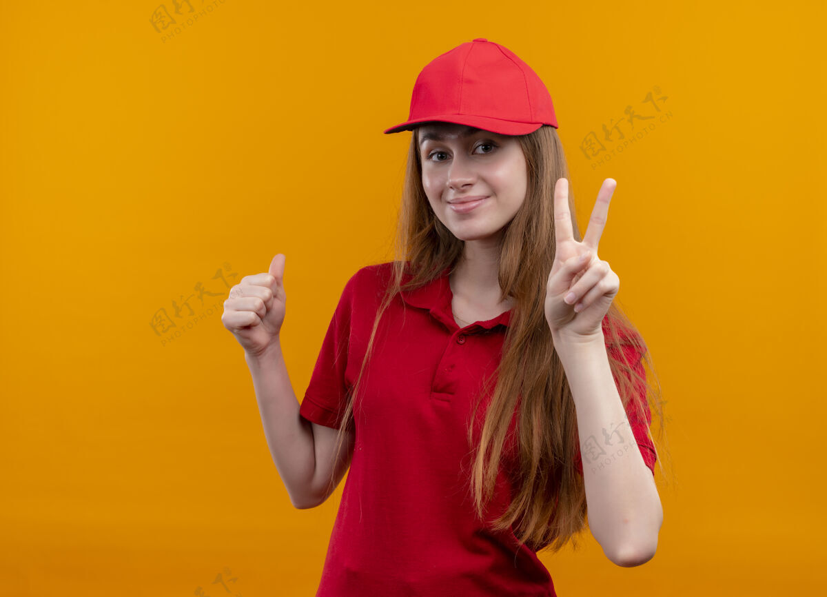 女孩穿着红色制服的年轻送货女孩高兴地做和平的标志 并在孤立的橙色空间与复制空间大拇指了空格橙色标志