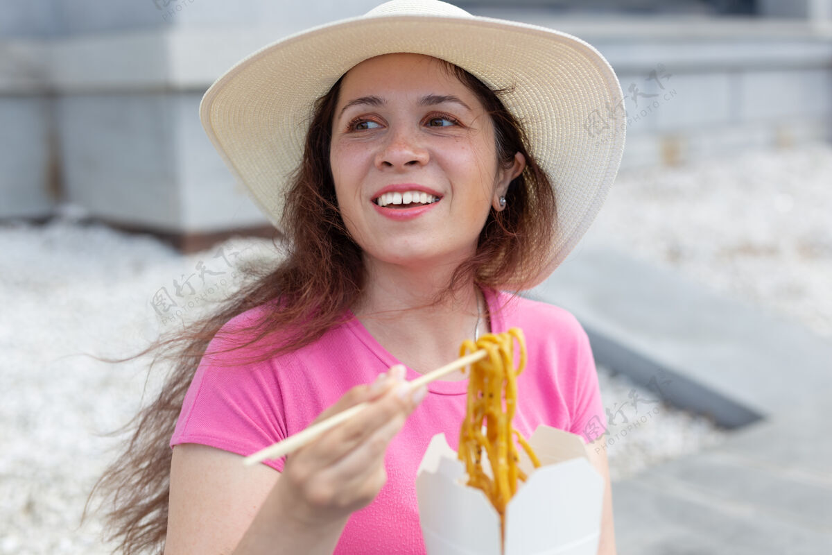 时尚年轻积极的女孩戴着帽子 用木筷吃中国面条作为外卖午餐的概念帽子魅力乌冬
