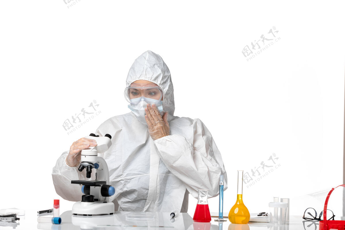 病毒正面图：女医生穿着防护服 戴着口罩 因为在白色办公桌上用显微镜观察病毒大流行 溅起病毒白色使用喷溅