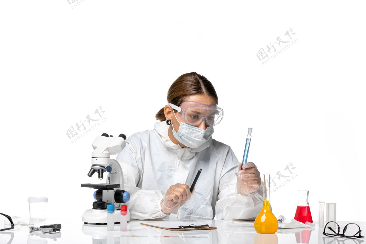 药品正面图：女医生穿着防护服 戴着口罩 拿着装有蓝色溶液的烧瓶 白色办公桌上有冠状病毒大流行医疗办公桌烧瓶