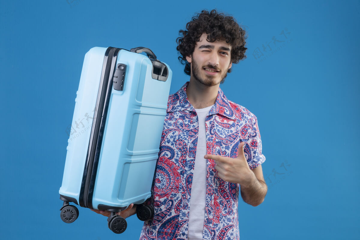 手提箱自信的年轻英俊的旅行家男人拿着手提箱指着它 在有复制空间的孤立的蓝色空间里眨眼男人拿着自信