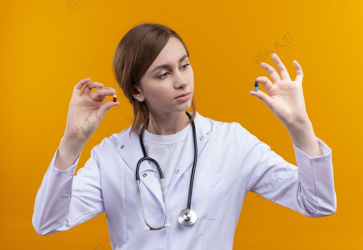 女性自信的年轻女医生穿着医用长袍和听诊器拿着医用胶囊 在孤立的橙色空间里看着它们信心医生年轻