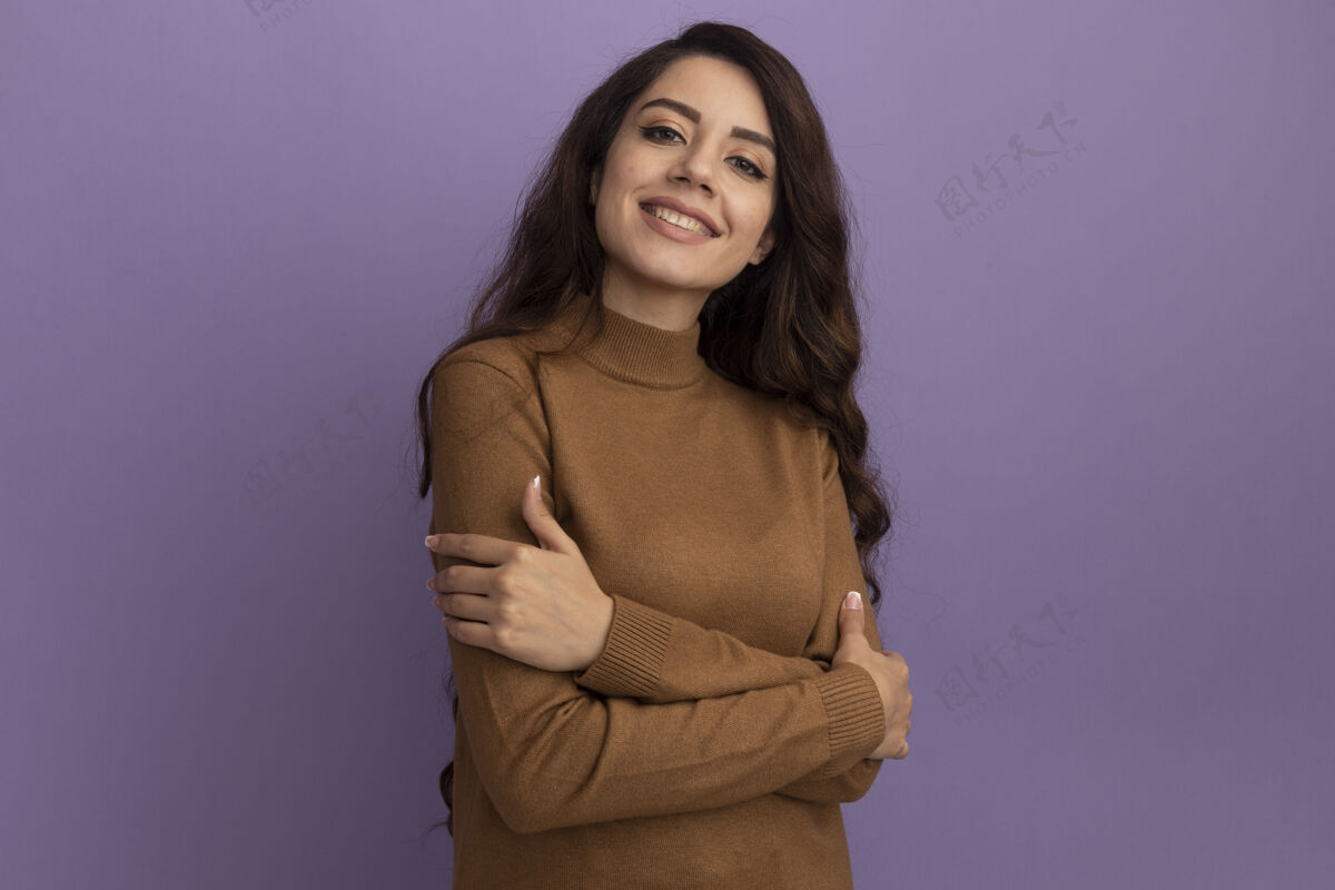 美丽带着微笑的年轻漂亮女孩穿着棕色高领毛衣双手交叉隔离在紫色的墙上 留着复制空间穿着高领毛衣脸