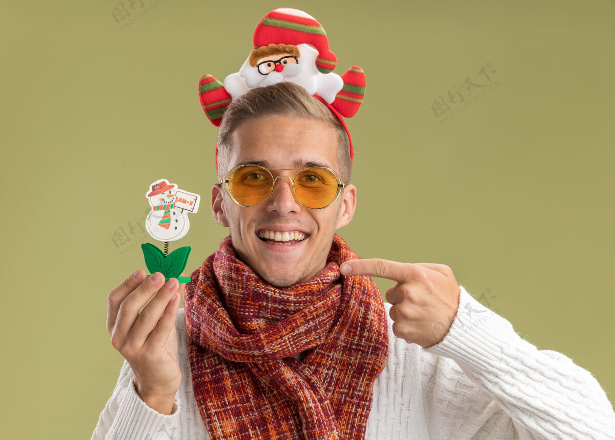围巾快乐的年轻帅哥戴着圣诞老人的头带和围巾看着相机拿着并指着橄榄绿背景上孤立的雪人玩具抱着快乐指着