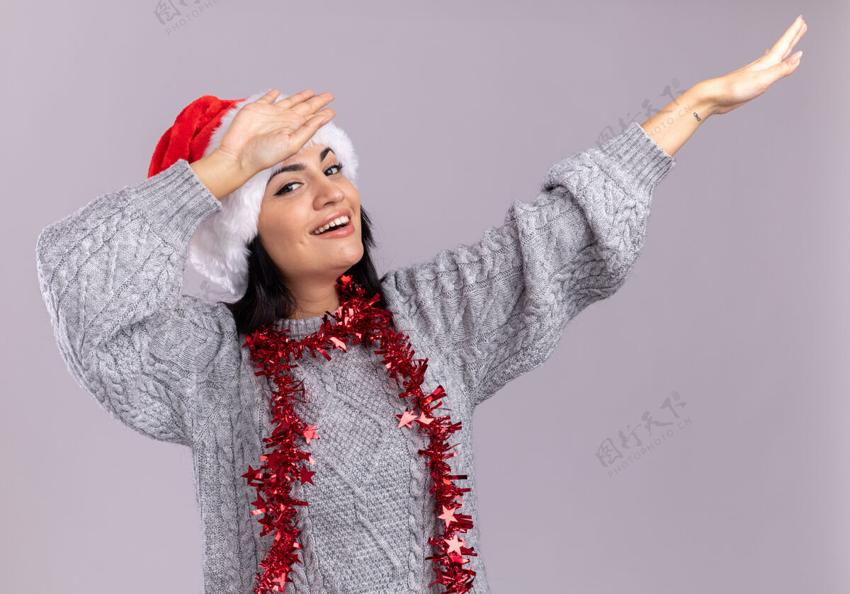 白色快乐的年轻白人女孩戴着圣诞帽 脖子上戴着金箔花环 看着相机在白色背景上做着一个孤立的手势花环轻拍手势