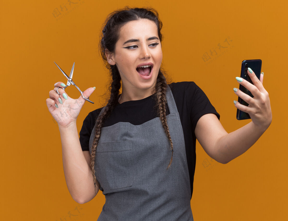 年轻人快乐的年轻女理发师穿着制服 手里拿着剪刀 看着孤立在橙色墙上的手机衣服剪刀站着