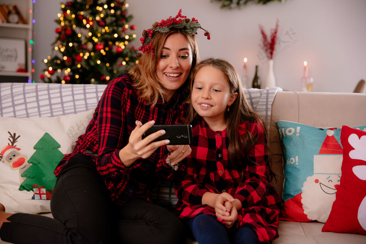 圣诞快乐快乐的妈妈和女儿坐在沙发上看电话 在家里享受圣诞节时光房子快乐电话