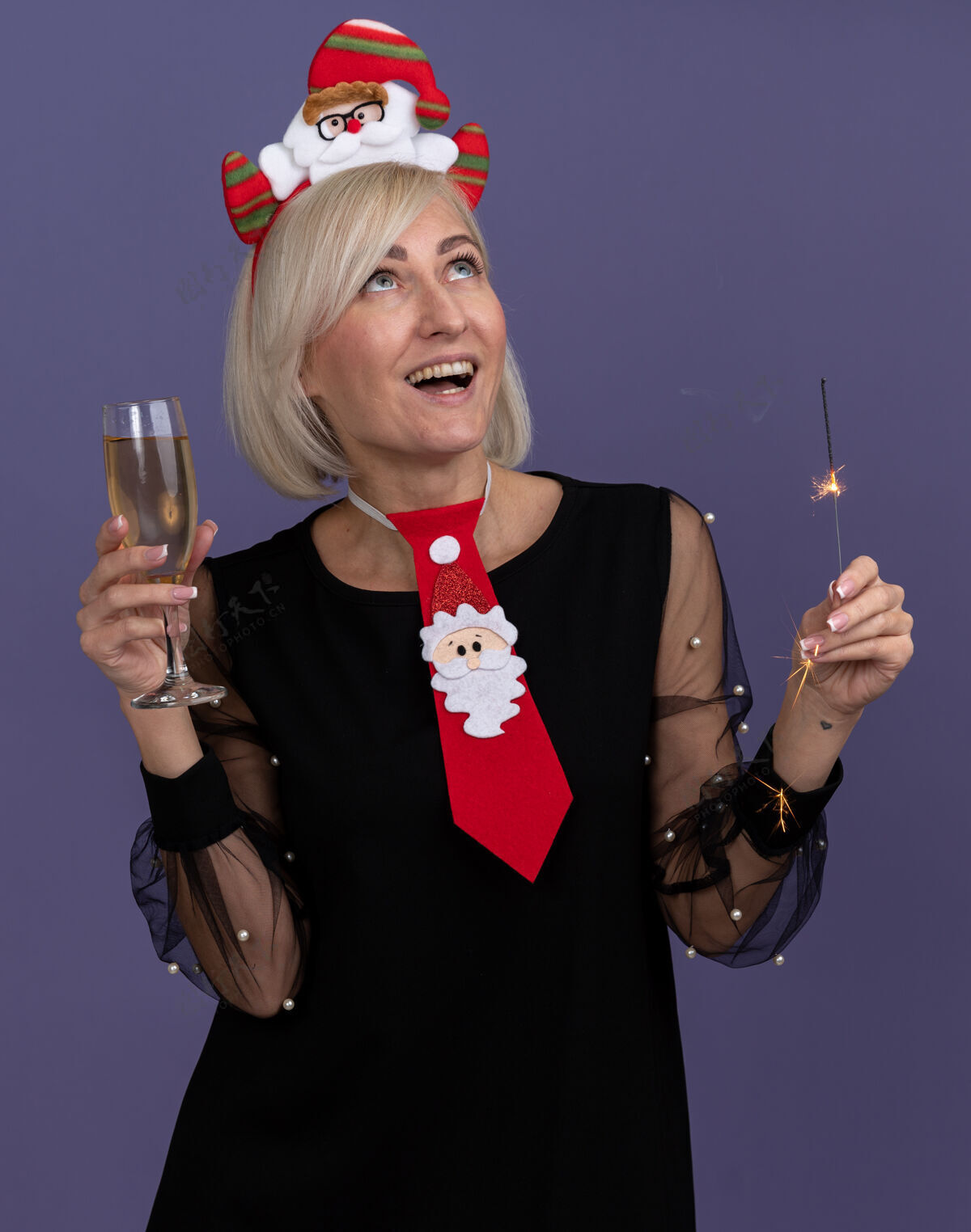 新年快乐快乐的中年金发女人戴着圣诞老人的头带 打着领带 手里拿着节日的火花灯和一杯香槟 在紫色的背景上抬头仰望孤立的人群头带圣诞快乐女人