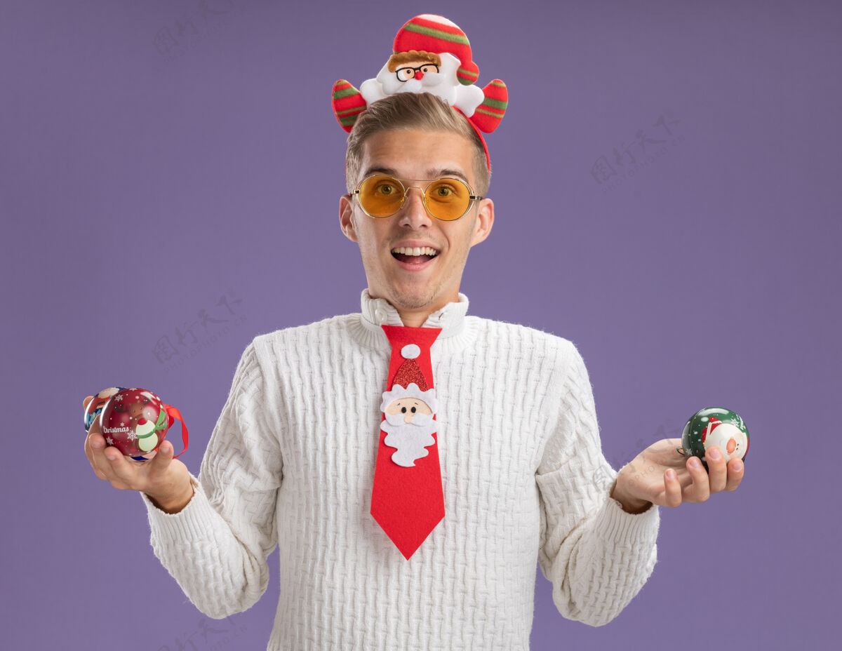圣诞老人印象深刻的年轻帅哥戴着圣诞老人的头带和领带 戴着眼镜 拿着圣诞球饰品 看着隔离在紫色背景上的相机眼镜圣诞老人圣诞快乐