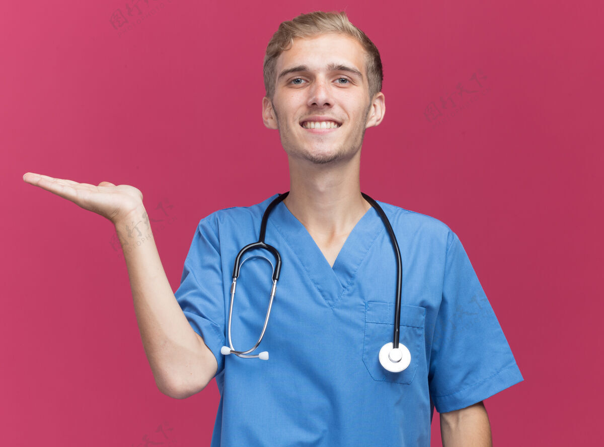 男性微笑着的年轻男医生 穿着医生制服 带听诊器 假装拿着隔离在粉色墙上的东西 留着复印空间制服人公民