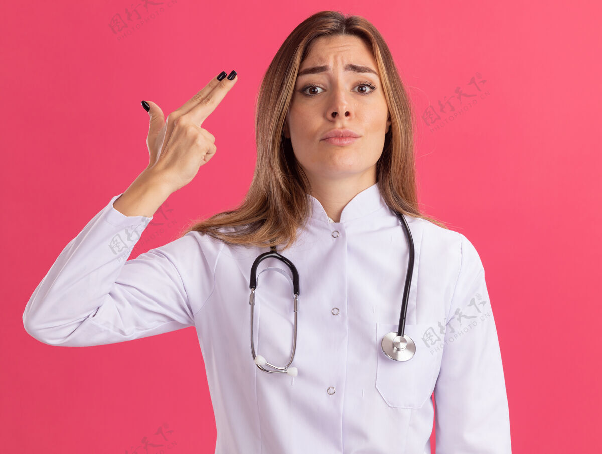 年轻人年轻的女医生穿着医用长袍 听诊器显示手枪的手势 隔离在粉红色的墙上手枪穿着感情