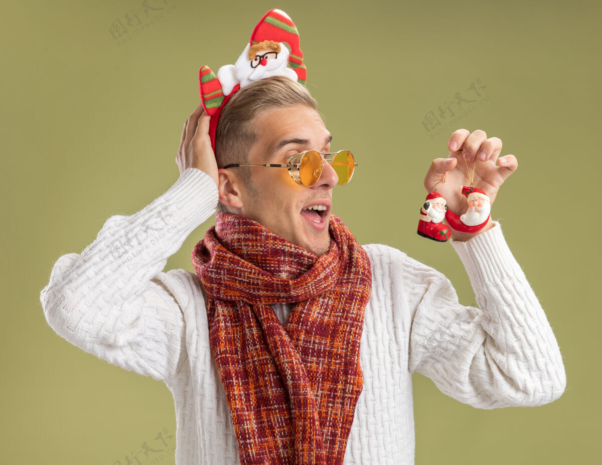 头带印象深刻的年轻帅哥戴着圣诞老人的头带和围巾拿着圣诞老人看圣诞饰品把手放在脑后橄榄绿的背景隔离圣诞老人装饰围巾