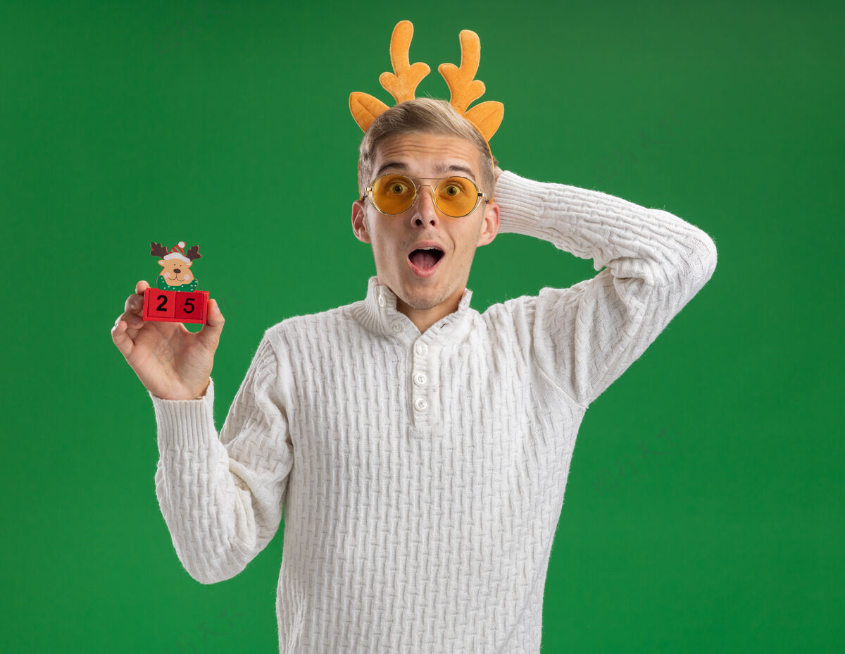 印象令人印象深刻的年轻帅哥戴着驯鹿鹿角头带戴着眼镜拿着圣诞树玩具和日期看着相机保持手在头后面隔离在绿色背景上头驯鹿绿色