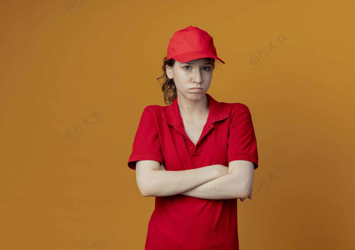 送货令人失望的年轻漂亮的送货女孩穿着红色制服 戴着帽子站着 封闭的姿势孤立在橙色背景上 留有复印空间红色橙色制服