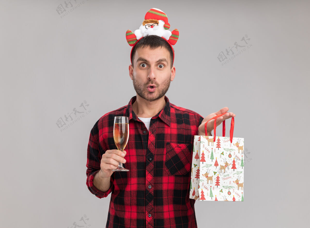 白色令人印象深刻的年轻白种人戴着圣诞头带拿着圣诞礼品袋和一杯香槟看着隔离在白色背景上的相机复制礼物圣诞快乐