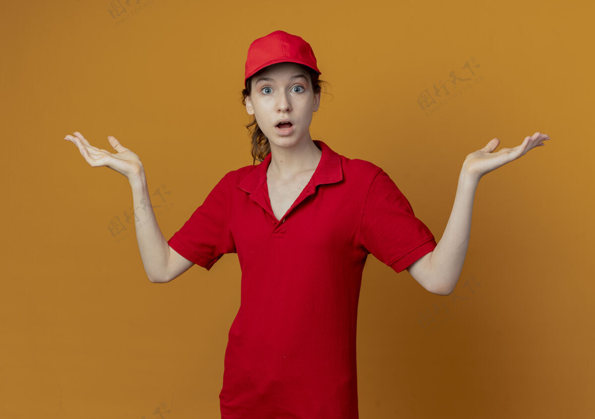 帽子令人印象深刻的年轻漂亮的送货女孩 穿着红色制服 戴着帽子 两手空空地看着橙色背景上孤立的摄像机女孩制服橙色