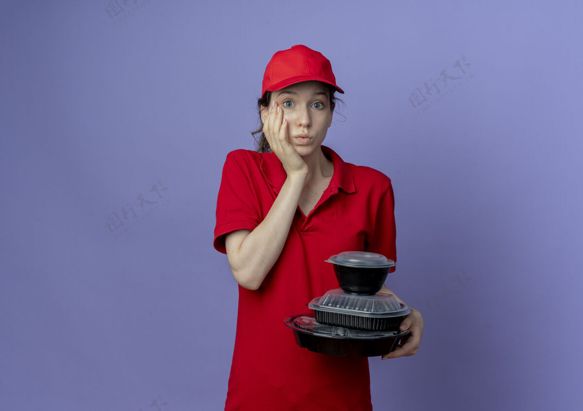 食物令人印象深刻的年轻漂亮的送货女孩穿着红色制服和帽子拿着食品容器把手放在脸上隔离在紫色背景与复制空间红色年轻手