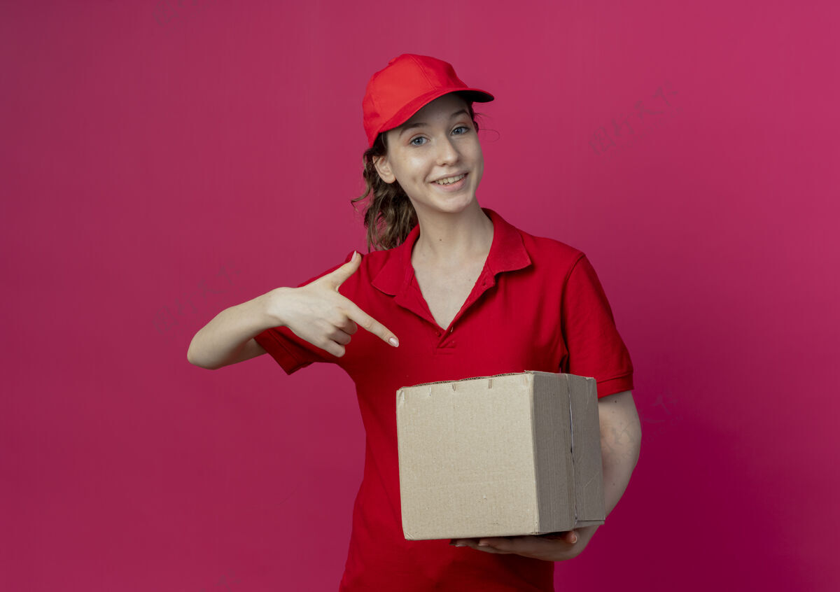 制服微笑着的年轻漂亮的送货女孩 穿着红色制服 戴着帽子 拿着并指着一个在深红色背景下有复印空间的纸箱纸箱指向盒子