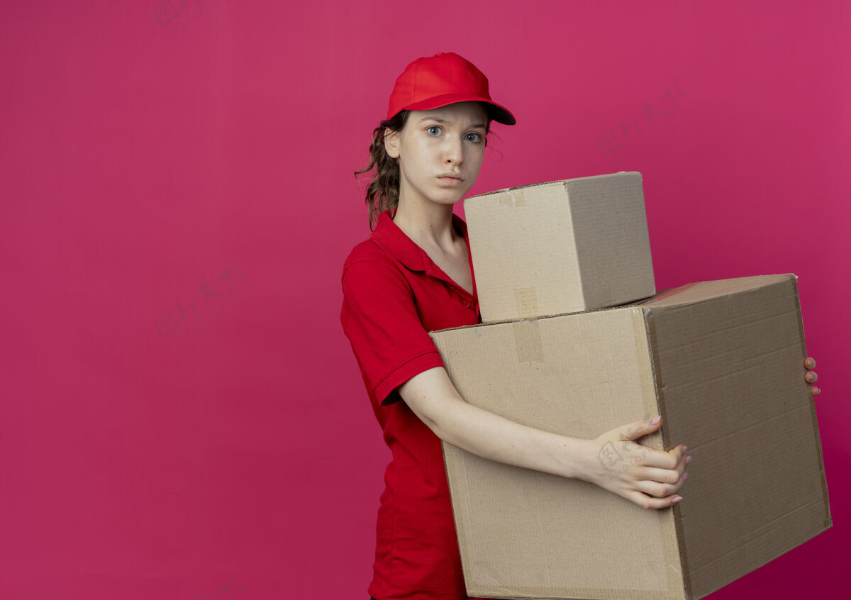 制服令人印象深刻的年轻漂亮的送货女孩在红色制服和帽子举行纸箱孤立的深红色背景与复制空间印象漂亮盒子