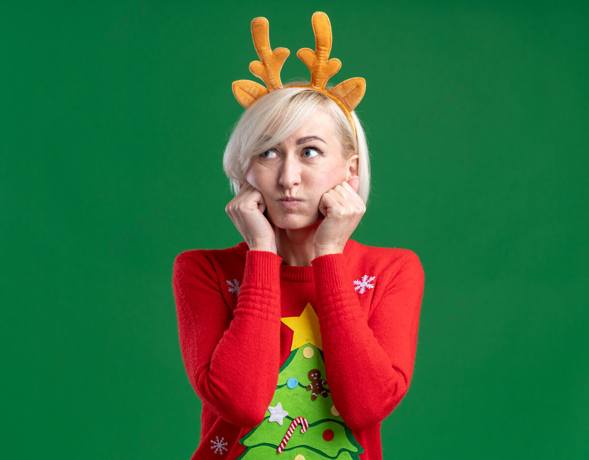 无聊无聊的中年金发女人戴着圣诞驯鹿鹿角头带和圣诞毛衣看着一边鼓起的脸颊保持双手在脸上绿色背景隔离圣诞节手圣诞快乐