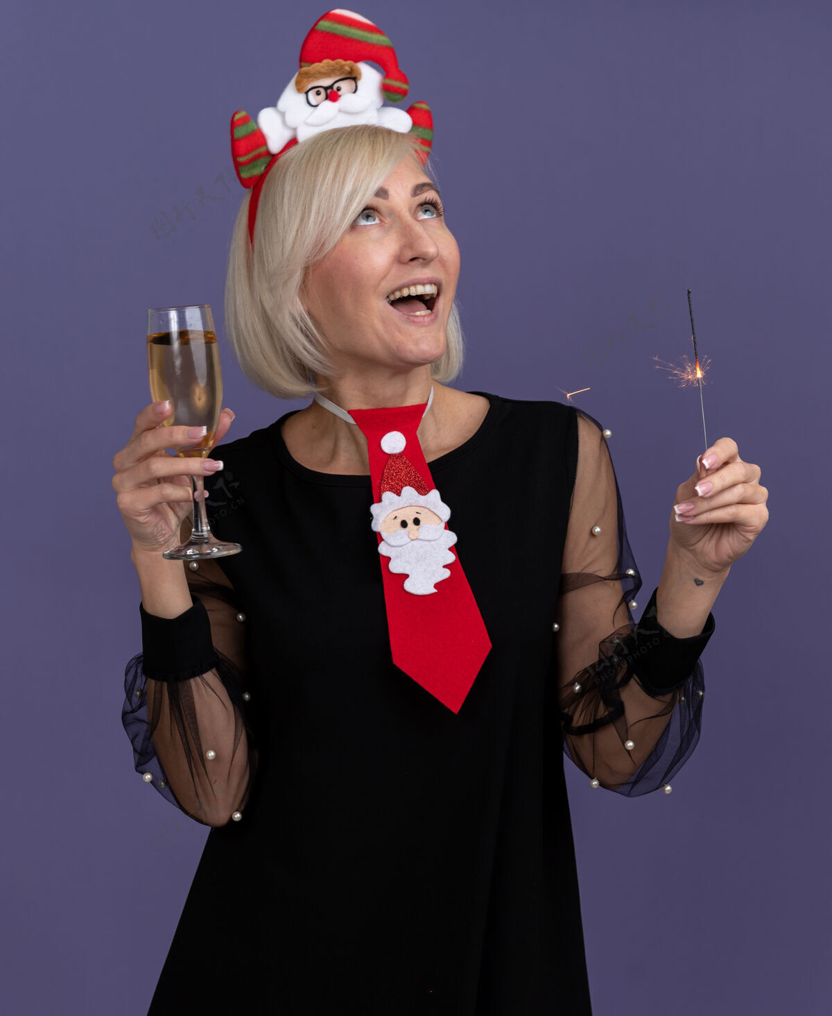 印象令人印象深刻的中年金发女人戴着圣诞老人的头带和领带 拿着节日火花和一杯香槟在紫色的背景上孤立地看着头带圣诞快乐女人