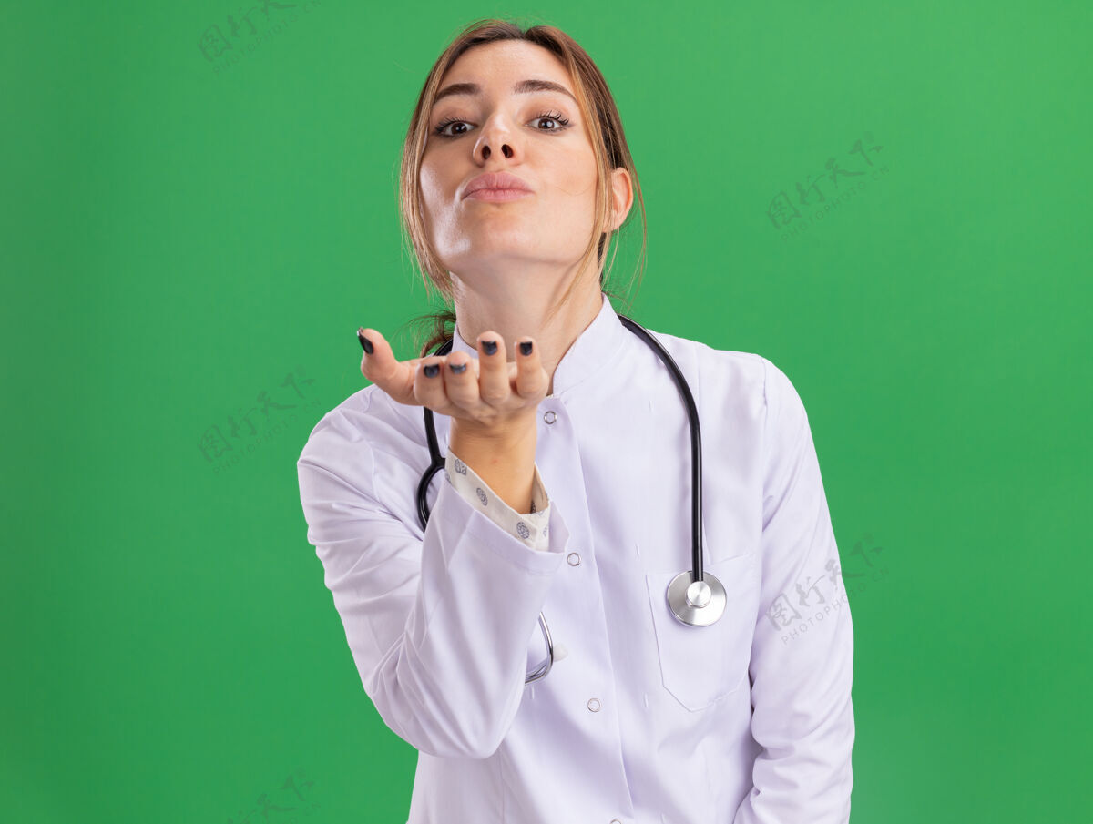 人高兴的年轻女医生穿着医用长袍 听诊器显示亲吻的手势隔离在绿色的墙上表演年轻人医学